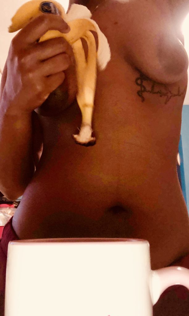Cara holding an open banana, naked from the waist up, with a coffee cup in the foreground in post titled Breakfast Treat 